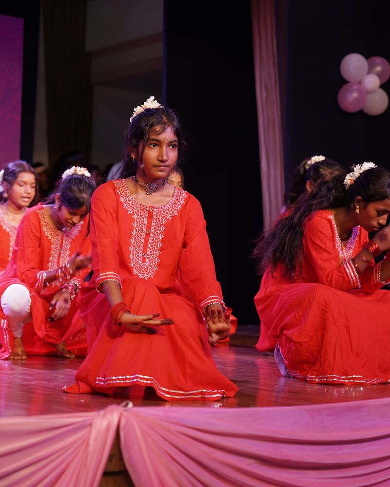 A girl in an orange kurta with jasmine in her hair, kneeling mid-dance at the front of the stage