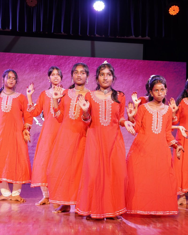 A row of girls in matching orange kurtas, palms out, dancing together under the stage lights