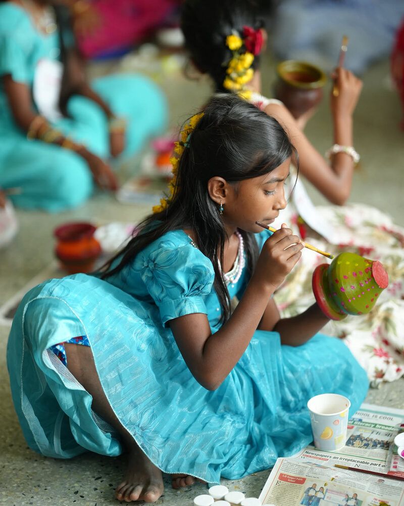 A girl in a blue dress painting a clay pot, surrounded by other children painting