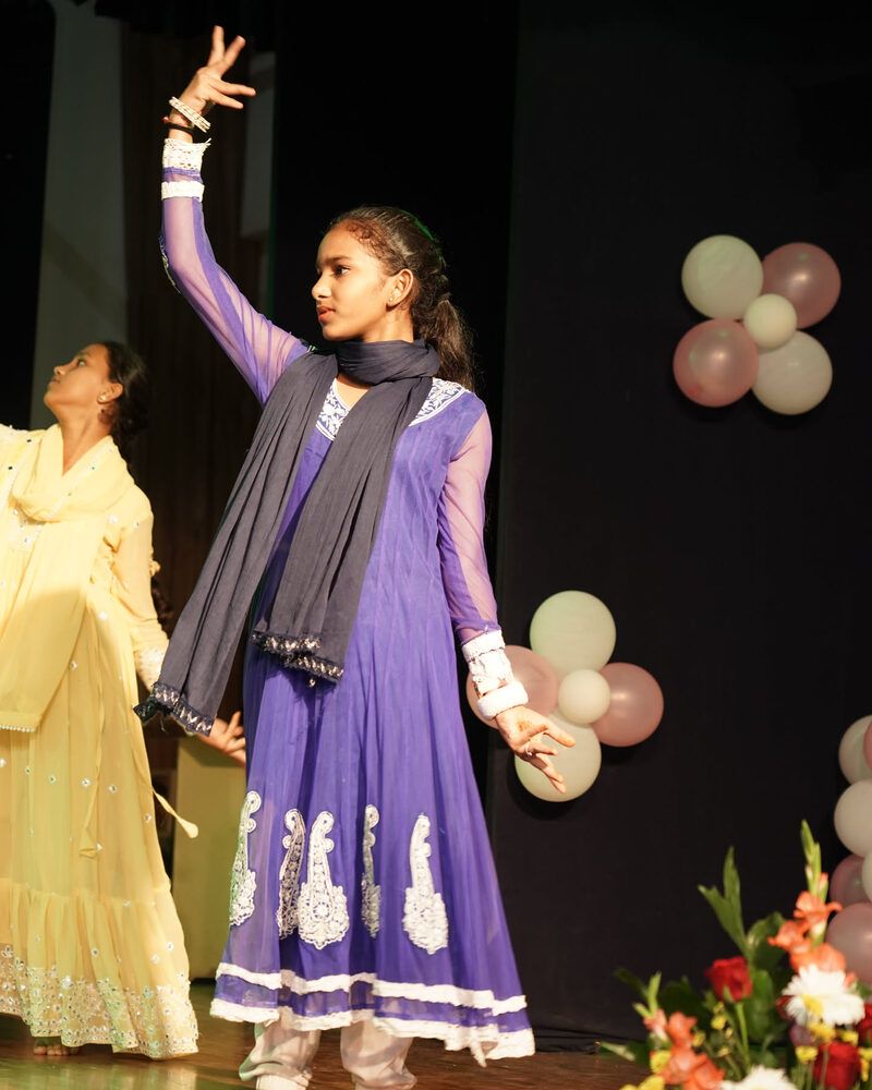 A girl in a purple kurta dancing a solo, one arm raised, beside a bank of flowers on stage
