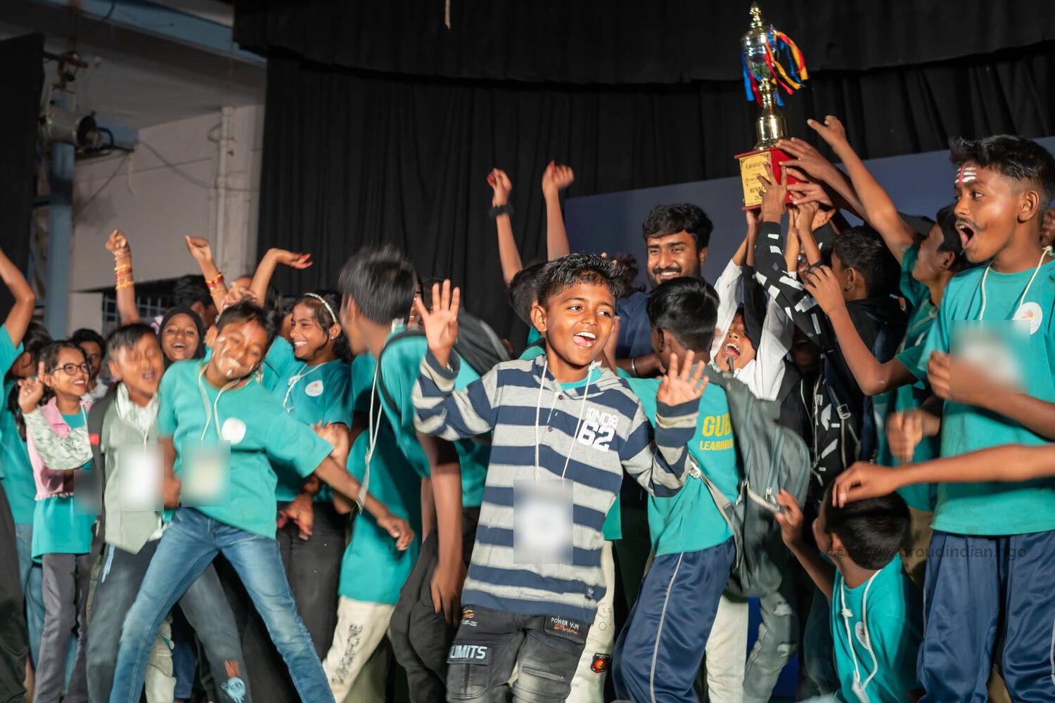 Children in teal T-shirts cheering as their team lifts a trophy on stage at Kalakriti 2.0