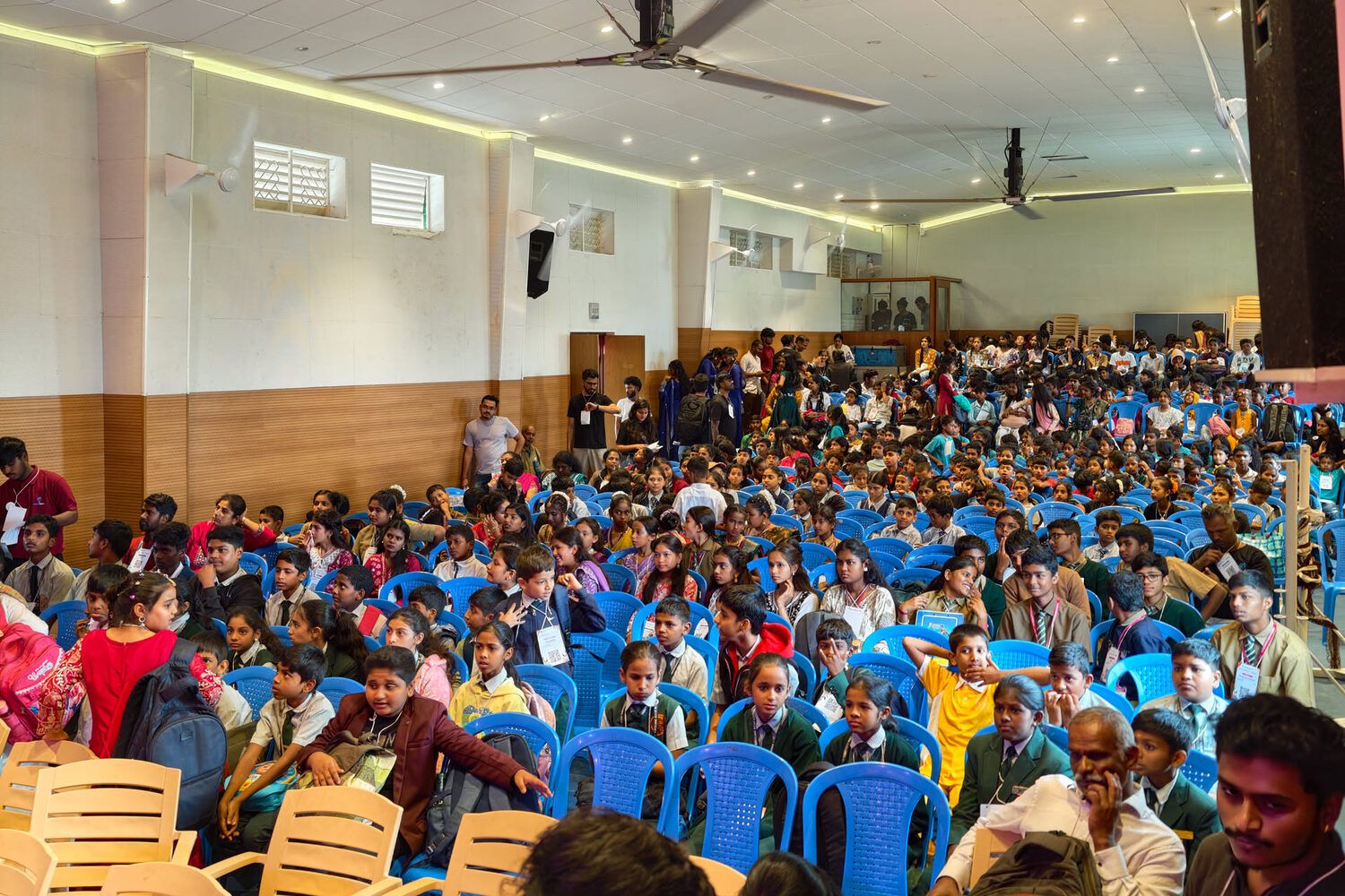 A hall packed with children on blue chairs, waiting for the stage events at Kalakriti 3.0